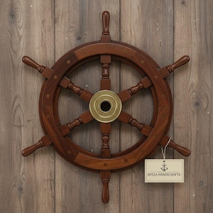 May include: A wooden ship's wheel with a brass center, mounted on a wooden wall. The wheel is dark brown with eight spokes and a brass center. A tag with the text "AYEZA HANDICRAFTS" hangs from the wheel.