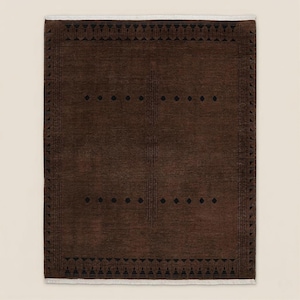 May include: A rectangular brown rug with a geometric design. The rug features a dark brown field with black diamond and line patterns. The border has a repeating pattern of triangles and other shapes. The rug has a white fringe on the short ends.