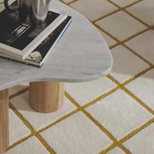 Puede incluir: Una escena de salón moderno con una mesa de centro con tapa de mármol y patas de madera. Una tetera plateada descansa sobre una pila de libros. La alfombra blanquecina tiene un patrón de cuadrícula con líneas doradas.