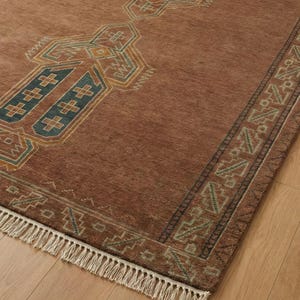 May include: A rectangular, rust-colored rug with an intricate geometric pattern in teal, gold, and navy blue. The rug features a central design and a border with a fringe along one edge. The rug is made of a woven material.