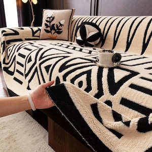 May include: A beige and black patterned sofa cover with matching throw pillows. The cover features a geometric design, and a hand is holding up a corner of the cover to show the texture. A cup of coffee sits on the sofa.