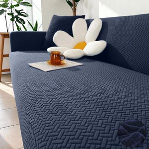 Könnte beinhalten: Marineblaue Sofahusse mit einem strukturierten geometrischen Muster. Das Sofa hat ein dunkelblaues Kissen und ein großes weißes kissenförmiges Kissen. Eine kleine Glastasse und eine Zeitschrift liegen auf dem Sofa.