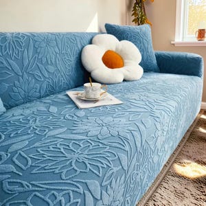 Puede incluir: Una funda de sofá azul claro con un estampado floral en relieve. Se ve un cojín a juego y un gran cojín con forma de flor. Una taza y un platillo descansan sobre una revista.
