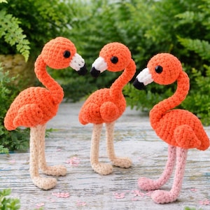 Pode incluir: Três flamingos de crochê feitos à mão. Cada flamingo tem um corpo laranja, um bico branco e preto e olhos pretos. As pernas são bege e rosa. O fundo é folhagem verde.