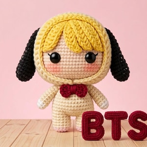 Puede incluir: Muñeca de ganchillo hecha a mano con una cubierta para la cabeza amarilla y negra, un cuerpo color crema y una pajarita roja. La muñeca tiene ojos negros grandes y mejillas rosadas. Las letras "BTS" están tejidas en rojo delante de la muñeca.