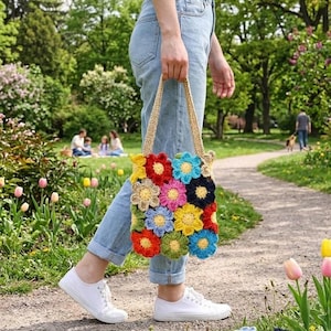 Pode incluir: Uma sacola tote de crochê com um design floral colorido. A sacola apresenta flores em tons de vermelho, amarelo, azul e laranja. Uma pessoa carrega a sacola enquanto caminha por um caminho em um parque.