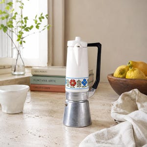 May include: A stovetop espresso maker with a white ceramic upper body featuring a colorful floral pattern of red and blue flowers. The base is made of silver-toned metal, and it includes a black handle and a white lid with a small knob.