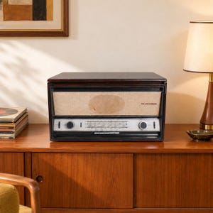 Może przedstawiać: Ciemnobrązowe drewniane radio Telefunken o prostokątnym kształcie, wyposażone w srebrny panel sterowania z dwoma okrągłymi pokrętłami i beżową maskownicą głośnika. Radio posiada szklany front i jest umieszczone na drewnianej powierzchni.