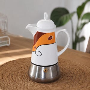 May include: A stovetop coffee maker with a white ceramic upper body featuring an orange and brown graphic design with coffee bean motifs. The base is made of polished silver-tone metal, and the lid has a small handle. It includes a side handle.