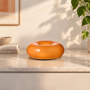 Op de afbeelding: Een glanzende, donutvormige oranje lamp met een glad, afgerond oppervlak en een centrale opening. De lamp is voorzien van een wit netsnoer dat aan de zijkant uitsteekt en rust op een vlak oppervlak, met een warme, reflecterende gloed.
