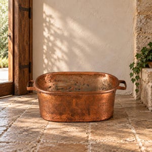 May include: An oval-shaped copper basin featuring a weathered, metallic finish with visible patina and oxidation. The basin includes two sturdy, flat metal handles attached to opposite sides, providing a functional grip for this open-top vessel.