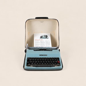 May include: A light blue Olivetti Lettera 32 typewriter housed in its original dark carrying case. The typewriter features a black QWERTY keyboard and includes an instructional manual with the text Olivetti Lettera 32 visible inside the case.