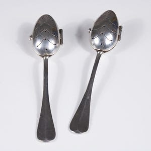 Op de afbeelding: Twee zilverkleurige metalen thee-eieren, elk voorzien van een scharnierende, eivormige kop met een decoratief chevronpatroon van kleine perforaties en een lange, slanke steel. De thee-eieren liggen naast elkaar en tonen hun metalen afwerking.