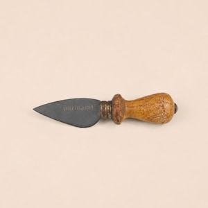 May include: A cheese knife featuring a dark gray, triangular metal blade with the word parmerat embossed on the surface. The handle is crafted from light brown, textured wood with a rounded end and a metallic ring connecting it to the blade.