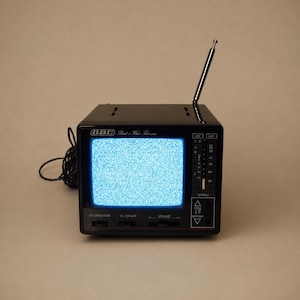 May include: A black GBC portable television with a glowing blue static screen. The device features a retractable silver antenna, a side-mounted channel selector, and front-facing controls labelled Interruttore, V.L, V.H, UHF, and Volume.