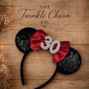 May include: Black sequined Minnie Mouse ears with a red sequined bow and a silver "30" in the center. The headband is black, and the background is a wooden surface. The text "The Twinkle Charm Co." is above the ears.