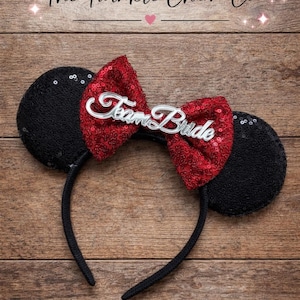 May include: Black sequined mouse ears with a red sequined bow featuring the text "Team Bride" in silver script. The headband is black. The image also includes the text "The Twinkle Charm Co."