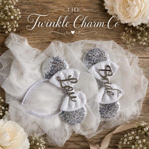 May include: Two white headbands featuring silver sequin ears and white fabric bows. One bow displays the silver script text "Love" and the other displays the silver script text "Bride". The headbands are placed on a white tulle fabric background.