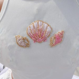 Può includere: Canotta bianca a coste ornata da tre disegni di conchiglie decorate con perline. Le conchiglie presentano perline rosa, bianche e dorate, creando un'estetica costiera. La canotta è un capo semplice e casual.
