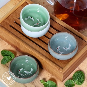 Puede incluir: Un juego de tres tazas de té de cerámica con un diseño de peces koi. Las tazas son en tonos verdes y azules, con un borde dorado. Las tazas están sobre una bandeja de madera, con una tetera en el fondo.