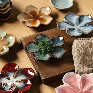 May include: A selection of ceramic flower-shaped dishes in shades of blue, red, and pink. Some dishes contain food, including seaweed and a brown liquid. The dishes are arranged on a wooden surface, with a neutral-coloured cloth visible.