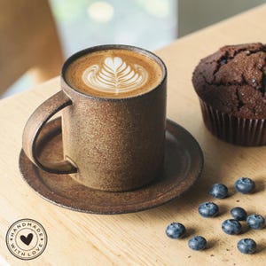 Puede incluir: Una taza de cerámica marrón con asa, llena de arte latte, se encuentra sobre un platillo a juego. Un muffin de chocolate negro y arándanos esparcidos están al lado de la taza. La taza tiene una pegatina "HANDMADE WITH LOVE".