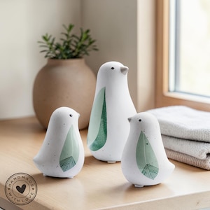 Può includere: Tre statuette di uccelli in ceramica bianca con accenti di foglie verdi e dettagli maculati. Gli uccelli sono di diverse dimensioni e sono disposti su una superficie di legno. L'uccello più piccolo ha il logo "HANDMADE WITH LOVE".