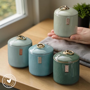Pode incluir: Quatro potes de cerâmica em tons de azul e verde com alças douradas e pequenas etiquetas retangulares. Os potes têm um acabamento craquelado. Um pote está sendo segurado. Os potes estão em uma superfície de madeira.