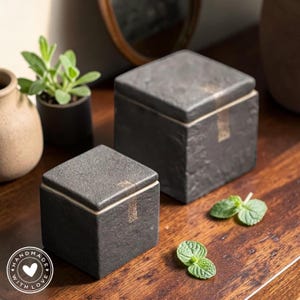 Op de afbeelding: Twee donkergrijze vierkante dozen met deksels, mogelijk voor opslag of decoratie. De dozen hebben een textuur en een goudkleurig accent op het deksel. De afbeelding bevat ook een kleine potplant, een vaas en een takje munt op een houten oppervlak. De woorden "HANDMADE WITH LOVE" staan in een cirkel.
