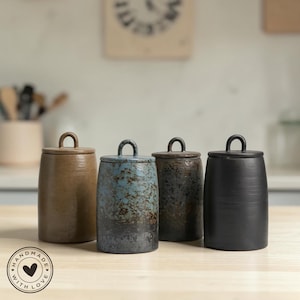 Könnte beinhalten: Vier handgefertigte Keramikgläser mit Deckeln. Die Gläser sind in Braun-, Blau- und Schwarztönen gehalten. Jedes Glas hat einen Schlaufengriff auf dem Deckel. Die Gläser stehen auf einer hellen Holzoberfläche.