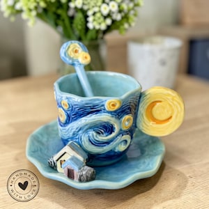Könnte beinhalten: Ein Keramik-Teebecher- und Untertassen-Set mit Löffel, inspiriert von Van Goghs "Sternennacht". Die blaue Tasse hat Wirbelmuster und gelbe Sterne. Der Griff ist ein gelber Halbmond. Eine kleine Hausfigur sitzt auf der Untertasse.