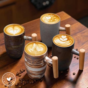 May include: Four ceramic coffee mugs with latte art, each with a unique design. The mugs have wooden handles and are filled with coffee. The mugs are brown, grey, and white, and are arranged on a wooden surface. The image includes the text "HANDMADE WITH LOVE".