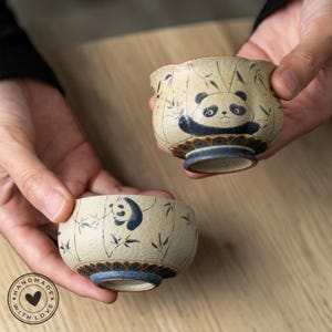 May include: Two handmade ceramic teacups with panda bear designs. The cups are beige with blue accents and feature hand-painted panda illustrations and bamboo stalks. The base of each cup has a blue ring. The cups are held in hands.