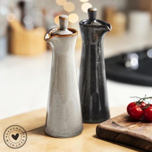 Puede incluir: Dos dispensadores de aceite de cerámica, uno gris claro y otro gris oscuro, con boquilla y tapón. Los dispensadores están sobre una superficie de madera, con una tabla de cortar de madera y tomates cherry en el fondo. La imagen incluye una pegatina "hecho a mano con amor".
