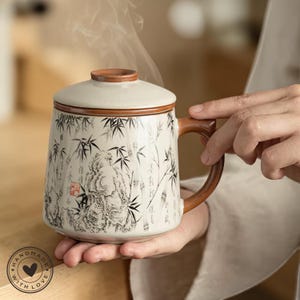 Può includere: Una tazza da tè in ceramica con coperchio, con un design a bambù e paesaggio in bianco e nero. La tazza ha un manico e un bordo marrone ed è tenuta da una persona. La tazza è etichettata "Handmade with Love".
