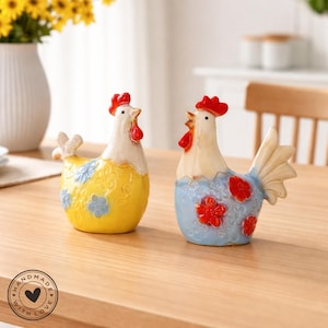Könnte beinhalten: Zwei Keramik-Hahnfiguren. Eine ist gelb mit blauen Blumenakzenten, die andere blau mit roten Blumenakzenten. Beide haben weiße Körper, rote Kämme und Kehllappen. Sie sind handgefertigt und dekorativ.