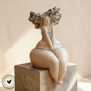 Peut inclure: Une sculpture en céramique d'une figure assise aux cheveux flottants. La figure est de couleur claire avec un motif de cœur sur la poitrine. La sculpture est assise sur un bloc de béton gris. Les mots "HANDMADE WITH LOVE" sont dans un cercle en bas à gauche.