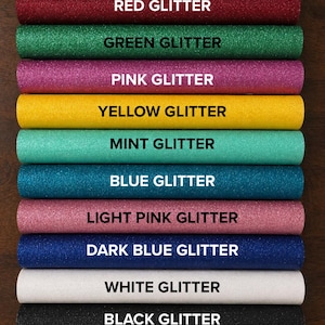 May include: A collection of twelve cylindrical rolls of glitter fabric arranged in a vertical stack. Each roll is labeled with its color: Silver, Red, Green, Pink, Yellow, Mint, Blue, Light Pink, Dark Blue, White, Black, and Gold Glitter.