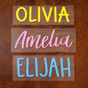 Pode incluir: Três decalques de nomes em vinil de transferência de calor. O decalque superior diz "OLIVIA" em amarelo, o decalque do meio diz "Amelia" em rosa e o decalque inferior diz "ELIJAH" em azul. Os decalques estão em um fundo transparente.