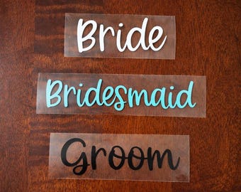 May include: Three clear rectangular decals with cursive text. The top decal reads "Bride" in white. The middle decal reads "Bridesmaid" in turquoise. The bottom decal reads "Groom" in black. The decals are on a dark wood surface.