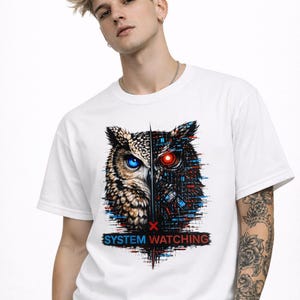 May include: White t-shirt featuring a split owl design. One side depicts a natural owl with a blue eye, the other a robotic owl with a red eye. The text "SYSTEM WATCHING" is below the graphic.