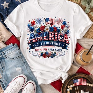 May include: White t-shirt with a patriotic design celebrating America's 250th birthday. The design includes the word "AMERICA" in red, white, and blue, along with flags, fireworks, and the dates July 4, 1776 - July 4, 2026. The shirt is paired with blue jeans and white sneakers.