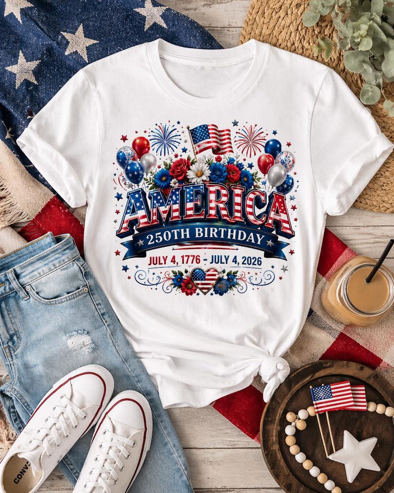 May include: White t-shirt with a patriotic design celebrating America's 250th birthday. The design includes the word "AMERICA" in red, white, and blue, along with flags, fireworks, and the dates July 4, 1776 - July 4, 2026. The shirt is paired with blue jeans and white sneakers.