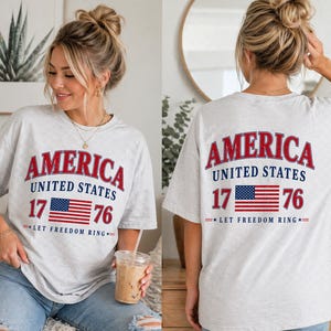Könnte beinhalten: Hellgraues T-Shirt mit dem Wort "AMERICA" in roter, weißer und blauer Schrift. Darunter stehen "UNITED STATES", die Zahl "17", eine Flagge, die Zahl "76" und der Satz "LET FREEDOM RING".