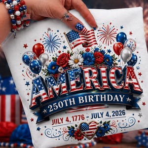 May include: White t-shirt with a patriotic design celebrating America's 250th birthday. The design features the word "AMERICA" in red, white, and blue, along with balloons, flowers, and the dates July 4, 1776 - July 4, 2026.