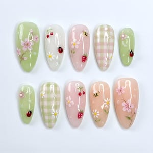 May include: A set of ten almond-shaped artificial nails with various designs. The nails feature hand-painted floral, fruit, and insect motifs, including cherries, strawberries, bees, and ladybugs, on a pastel color palette of green, white, and pink.