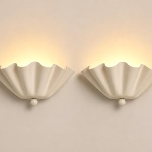 May include: A pair of off-white wall sconces featuring a pleated, fan-like shell shape with a small spherical accent at the base. Each fixture emits a warm, soft glow from the top, casting light upward against the wall surface in a uniform manner.