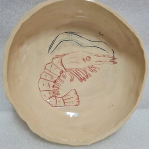 Könnte beinhalten: Handgefertigte Keramikschale mit cremefarbener Glasur. In der Mitte befindet sich eine rote Garnelenillustration und darüber eine schwarze Strichzeichnung. Der Rand der Schale hat eine unregelmäßige, organische Form.