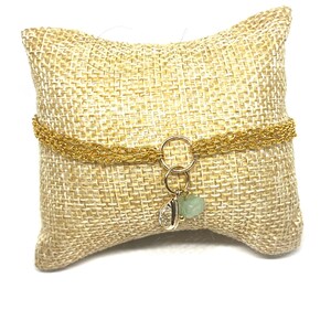 Op de afbeelding: Een goudkleurige kettingarmband met een kleine bedel met een lichtgroene steen en een zilverkleurig blad. De armband wordt gepresenteerd op een beige geweven kussen, waardoor een neutrale achtergrond ontstaat.
