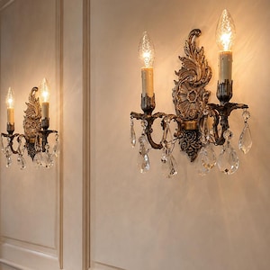 May include: Two ornate wall sconces featuring a dark metallic finish, intricate scrollwork, and hanging clear crystal teardrop prisms. Each sconce holds two candle-style light bulbs that emit a warm glow, mounted on a paneled wall above a wooden table.
